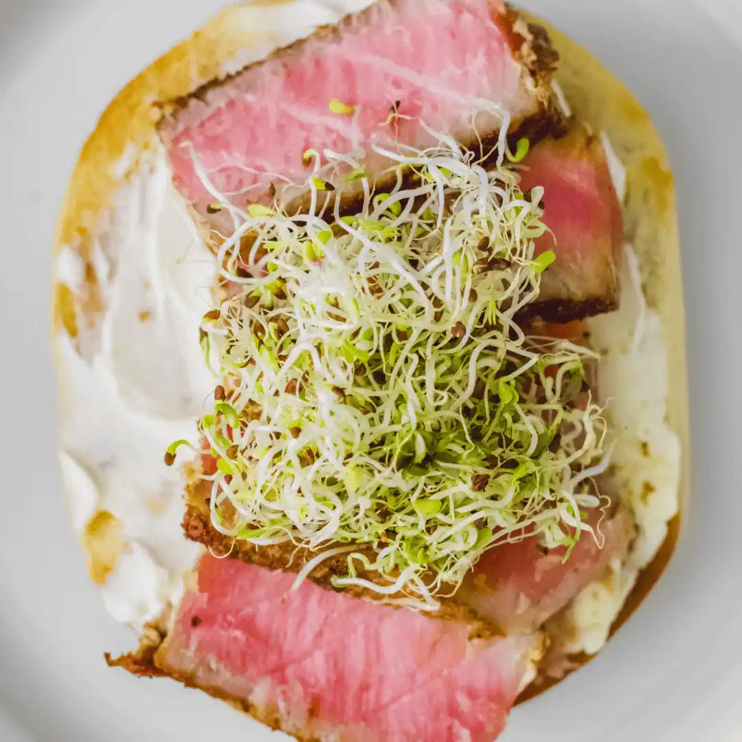 sliced seared steak on a toasted bun with mayo topped with fresh broccoli sprouts
